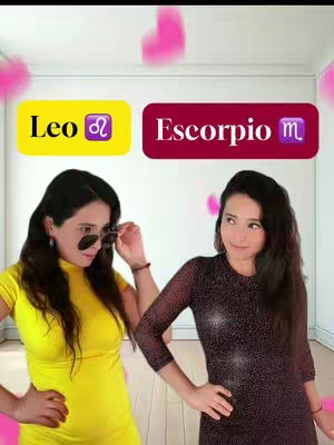 The image shows two women in a well-lit room or studio. One, on the left, wearing a yellow top and sunglasses, is holding up her sunglasses. The other, on the right, is wearing a dark dress with sparkling patterns, posing with one hand on the hip. Above them, slightly blurred pink hearts appear. There are two text boxes: one, yellow, with the word “Leo”, and the other, red, with the word “Escorpio”
