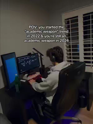A dimly lit image of a person sitting at a desk with a computer setup. The main focus is on the person, viewed from the back, wearing headphones and working at the computer. On the screen, mathematical equations are visible. The background elements include a window with blinds.