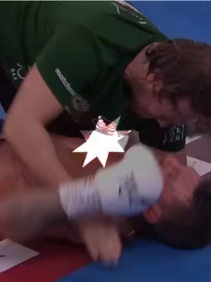 A close-up shot of a boxing match with two figures engaged. One person, wearing a green shirt, appears to be kneeling over another with white boxing gloves, who is lying on the ground. The image has a star-shaped censor covering a portion of the scene, and the background is a blue boxing ring mat.