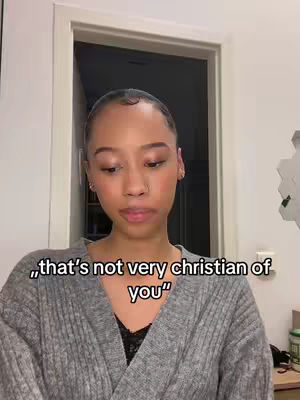 A medium shot shows a young woman with light skin, brown eyes, and dark hair pulled back. She faces the camera with a serious expression, lips slightly pursed. She wears a gray sweater over a black shirt. The background includes a partially visible white door frame.