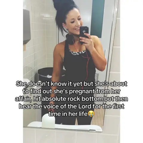 A woman takes a mirror selfie in a public restroom, smiling and holding a phone. She is wearing a black tank top and has her hair up in a bun with headphones around her neck. Text overlays are placed on the image.