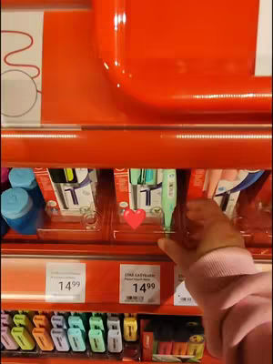 The cover image shows a close-up of a shopping shelf with pens on display. A hand wearing a pink sleeve is seen reaching for one of the green pens with a red heart overlayed. The shelves are in bright orange.