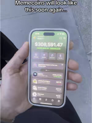 A hand holding a smartphone displaying a crypto wallet interface. The phone screen shows a balance of $308,591.47. The background appears blurred, possibly outdoors.