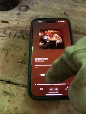 A person is holding a smartphone with a black case. The screen displays music playing from the app. The song "Nothing's New" by Rip Romeo is visible. The person's hand and part of their arm, wearing a green sleeve, is visible in the foreground.