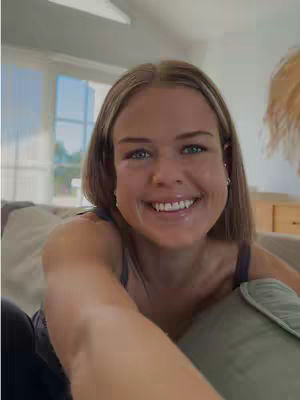 A selfie-style shot of a young woman with light brown hair smiling at the camera with a light filled background. The woman is in the foreground, her face and upper body taking up most of the frame. Her smile is wide, showing her white teeth, and her eyes are bright. She is holding the camera, and sitting in an area with natural light.