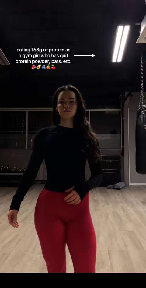 A woman stands in a gym setting, wearing a black long-sleeve shirt and red leggings. Text overlay indicates a focus on protein intake. The background includes gym equipment, such as a punching bag.