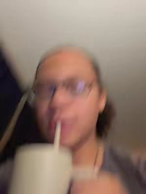 A blurry, somewhat out-of-focus selfie. The main subject is a person wearing glasses, holding a cup with a straw up to their mouth, appearing to be drinking. The background is dimly lit and indistinct, with a blurry white ceiling visible above.