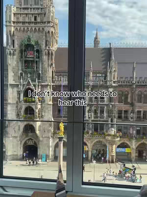 A view through a window of a large European building, likely a cathedral or town hall, with intricate architecture. The foreground consists of a window frame and a section of what appears to be the interior of a room. The background shows a bustling city square with people walking around. Added text overlayed on image.