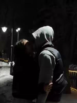 A nighttime scene with a couple embracing in a dimly lit, potentially outdoor setting. The man, facing away, is wearing a hooded jacket and vest, while the woman is positioned to the side, also in dark attire. Streetlights illuminate the background, highlighting the couple's intimate moment.