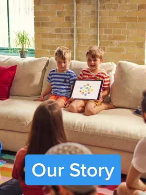 Two young boys are sitting on a cream-colored couch holding a tablet. A person's back is to the camera in the foreground, and the background features a brick wall and a window letting in light. The composition is balanced, with subjects in the foreground and background.