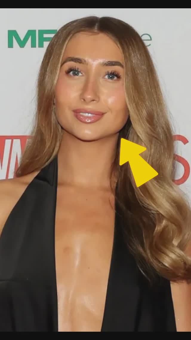 A headshot of a fair-skinned woman with long blonde hair and blue eyes, wearing a black top. She has a neutral expression, and the background is blurred. A yellow arrow points towards her jawline.