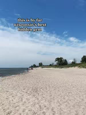 A wide shot of a sandy beach under a blue sky. The beach is in the foreground, with the ocean on the left and a grassy area with trees on the right. The sky is a bright blue with some fluffy white clouds. Text overlayed on the image says 'this is by far wisconsin's best hidden gem'.