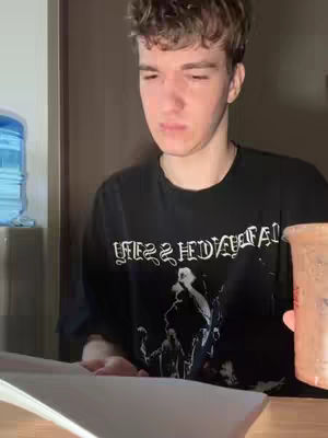 A young man with light skin is centered in the image, looking slightly downwards with a contemplative expression. He has short, tousled brown hair. He wears a black t-shirt with white text on it, and is holding a clear plastic cup in one hand while resting the other on a white paper in front of him on the desktop. Part of a water dispenser in the background is visible.
