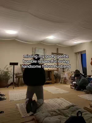 A person wearing grey pants is kneeling on a rug, positioned in the center of the frame. The background shows a room with wooden floors, white walls with a textured ceiling, and a cross on the wall.