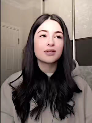 A woman with long, dark hair and a neutral expression is centered in the frame. She's wearing a light-colored hooded sweatshirt. The background includes a door and a mirrored closet.