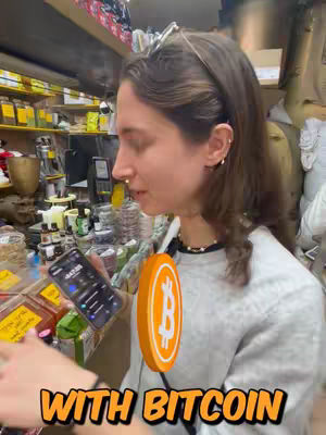 A woman stands in a store, holding a smartphone showing a cryptocurrency app. A large, animated Bitcoin logo is overlaid on the image. Text below the woman reads, 'WITH BITCOIN'. The scene has a brightly lit, commercial feel.