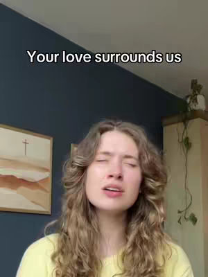 A woman with long wavy blonde hair is singing with her eyes closed in front of a dark blue wall. Above her, text reads 'Your love surrounds us.' To the left of the woman is a framed painting of an abstract cross and landscape. The overall tone is spiritual and emotive.