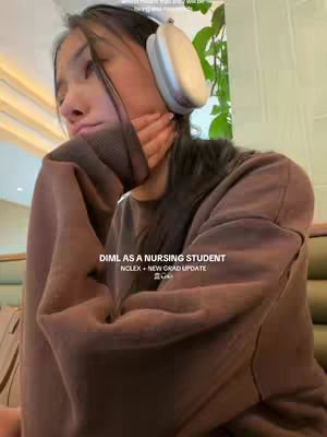 A young woman with long dark hair, wearing a brown sweater and white headphones, is looking to her left with a somewhat downcast expression. She is resting her head on her hand. The background is a soft beige color with glimpses of green plant life. Text overlays read 'DIML AS A NURSING STUDENT' and 'NCLEX + NEW GRAD UPDATE'.