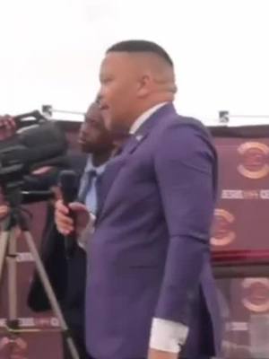 A man in a purple suit sings into a microphone, standing slightly off-center. Behind him, a man in a dark suit and tie stands out of focus, and there's a video camera on a tripod to the left. The overall image is well-lit, suggesting an event being recorded.