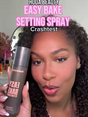 The cover image showcases a woman holding a bottle of Huda Beauty Easy Bake Setting Spray. The woman, with curly black hair, is captured in a close-up shot, with the product prominently displayed. The background is soft, likely of a room with hints of products and a mirror. Overlaid text in the pink color at the top reads 'HUDA BEAUTY EASY BAKE SETTING SPRAY Crashtest'.