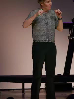 A woman with short blonde hair stands on a stage, dressed in a gray patterned button-down shirt and black pants. Her arms are raised, possibly gesturing, against a light pink background.