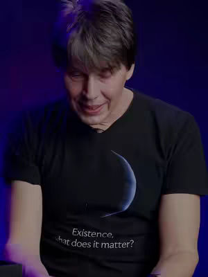 A man looks down towards the center of the frame, likely speaking. He's wearing a black v-neck t-shirt with an image of a crescent moon and the words, 'Existence, what does it matter?' written below. The background is a dark blue gradient. Portrait shot.