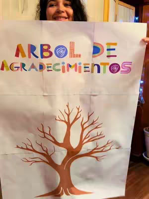 A woman holding up a white poster with a colorful text overlay and a drawing of a tree. The woman is smiling, and the background is a warmly lit room.