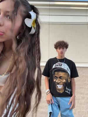 A young woman with long, wavy brown hair, partially obscuring the left side of the frame. She wears a white top and a gold necklace. The right side features a young man wearing a black t-shirt with a graphic of a smiling man with a basketball cap. He's in front of a beige, textured wall.