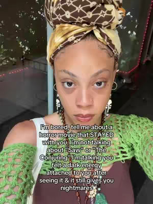 A selfie-style shot featuring a woman with a patterned head wrap, earrings, and a necklace. She's wearing a green crochet top over a brown garment. Text overlay adds context related to horror movies.