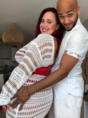 A man and a woman are embracing, appearing close and affectionate. The woman is on the left, wearing a white crocheted dress over a red top. The man on the right is in a white shirt and embraces her. They are both smiling at the camera in a warm and inviting setting. Lighting appears soft and diffused.