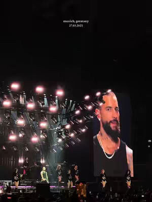 The image shows a live concert scene at night. The stage is illuminated with bright lights and filled with performers. In the foreground, several female dancers in black outfits are visible. On a large screen in the background, a close-up image of a man with a shaved head and a beard is displayed. The scene is energetic and visually striking.