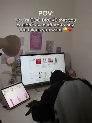 A person is seated at a desk with a laptop and a tablet. The laptop screen shows a website with product listings. The tablet screen is also displaying a web page with a similar look. The person is partially visible, wearing a black top. The setting is a well-lit room with items like a desk lamp, flowers, and wall decor. The overall composition is focused on the technology and the task at hand.