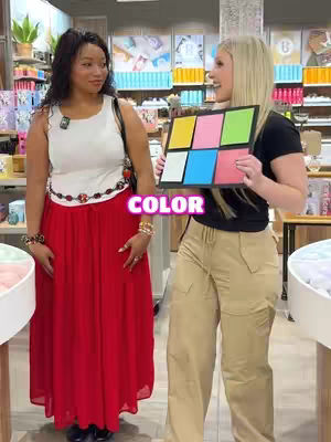 Two women stand in what appears to be a store, one holding up a color palette. The woman on the left wears a red maxi skirt and white top. The woman on the right wears a black top and tan pants and is holding a color palette.