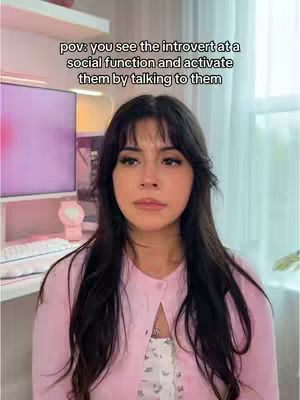The image shows a young woman indoors, likely in her room, with a soft, warm lighting. The background includes hints of a computer setup and a bright window, possibly indicating a workspace or bedroom. The woman has long, dark hair with bangs, a pink cardigan, and a white top. She has a neutral expression. The text overlay says: "pov: you see the introvert at a social function and activate them by talking to them".