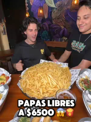 The cover image shows two young men posing with a massive platter of french fries. The fries are the focal point, overflowing from a metal serving dish. The hook text "PAPAS LIBRES A $6500" is prominently displayed. The background is slightly out of focus, suggesting a bar setting with patrons and dim lighting.