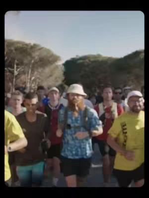 A group of people running in an outdoor setting. The focus is on a man with a beard wearing a blue shirt, hat, and backpack, surrounded by other runners. The general color palette is natural, with greens from trees and the blue sky visible. The runners are in various athletic attire, suggesting a race or a casual run.