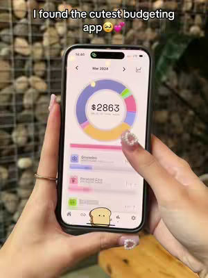 A person is holding a smartphone displaying a budgeting app interface. The app shows a pie chart representing financial allocations, the screen features a mix of pastel colors like purple, blue, and yellow, suggesting an aesthetic appeal. The person's hands are visible, adorned with polished nails and a gold ring. The background appears to be a neutral tone.