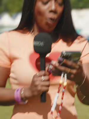 A woman with long black hair stands outdoors, with a microphone in one hand and a phone in the other. The image is slightly blurred.