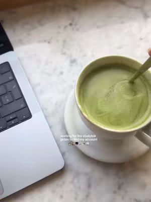 The image shows a top-down view of a laptop and a cup of matcha tea on a marble surface. A spoon is in the tea, and the laptop is open with the keyboard facing the viewer. The overall composition is light and airy.