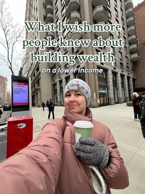 A selfie-style shot with the creator in the foreground holding a coffee cup, set against an urban background. The creator wears a grey beanie, a large dark pink jacket, and grey gloves. The background features a tall building and a street scene with pedestrians.