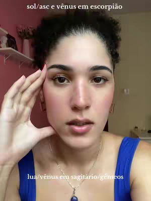 A close-up shot of a woman with light skin is framed from the chest up. She's looking directly at the camera with a neutral expression, her right hand gently touching her temple. She has dark, curly hair pulled back over her head. The background is softly blurred and shows a simple interior setting. The image has a natural lighting and a warm color palette, with hints of pink and blue.