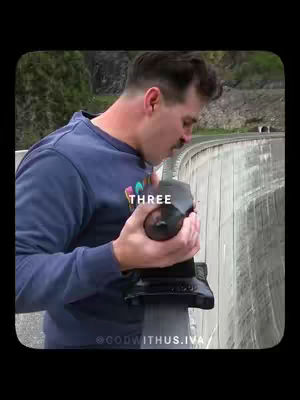 A man holding an object in his hands, stands in front of a dam. The word "THREE" is overlaid on the image.