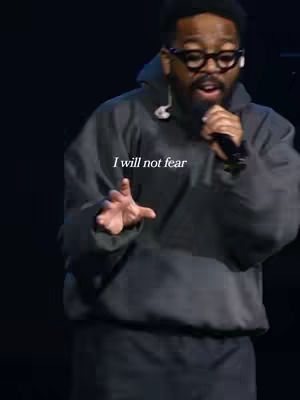 A man wearing glasses and a black hoodie is on stage in front of a dark background and holding a microphone while presenting with an open palm gesture. Overlay text reads, "I will not fear".