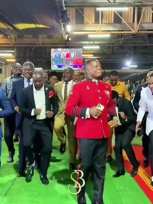 A group of formally dressed men, most in suits, are walking forward in a brightly lit indoor setting, likely a large hall. The central figure, dressed in a red jacket with black pants is holding a phone and leading the procession. The background is blurred, with a screen visible. A golden monogram logo is at the bottom.