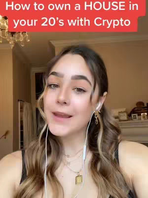 A woman with long, wavy brown and blonde hair is centered in the frame, wearing a black top. She smiles as she speaks, with a red rectangular text overlay at the top reading, "How to own a HOUSE in your 20's with Crypto." She has earbuds in, and a necklace with a letter pendant. The background includes a chandelier and glimpse of a room, with a soft, warm lighting.