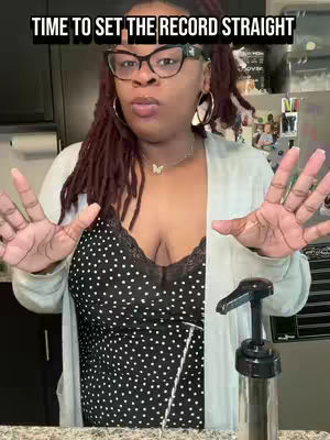 A woman with dark hair and glasses is centered in the frame, gesturing with her hands open. A black text overlay with white font is present above her head. She wears earrings and a patterned top over a black bra. A pump bottle is visible in the foreground on the right.