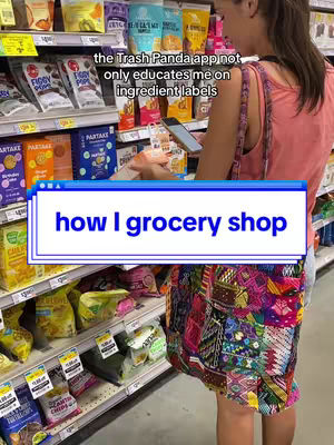 A person wearing a patterned skirt stands in a grocery store aisle, examining their phone. The scene is shot from behind the individual, framed to include the surrounding shelves stocked with various packaged foods. A large text overlay reading 'how I grocery shop' is prominently featured in the foreground. A text is overlaid stating "the Trash Panda app not only educates me on ingredient labels". The layout uses natural light, emphasizing the products and person.