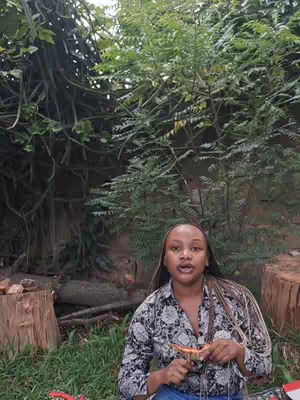 A woman is sitting in a natural setting with green foliage in the background and a wooden stump on the right. The woman has her mouth open slightly and she is holding a small object in her hand. She is wearing a patterned shirt with a floral design.