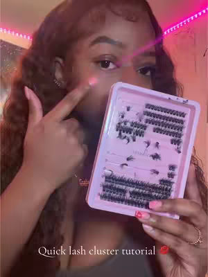 A woman with long brown wavy hair is holding a pink lash tray in front of her face. She is pointing to her cheek with her index finger. Text "Quick lash cluster tutorial" is at the bottom. The background has a pink light.