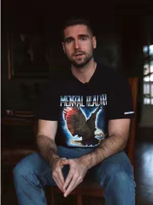 A man wearing a black t-shirt with a design of an eagle, sitting in a chair with his hands clasped. The background is dark and indistinct.